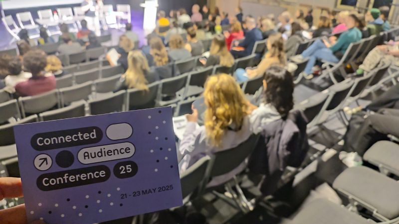 Vorne scharf gestellt das Konferenzprogramm, im Hintergrund unscharf die Reihen des Publikums.