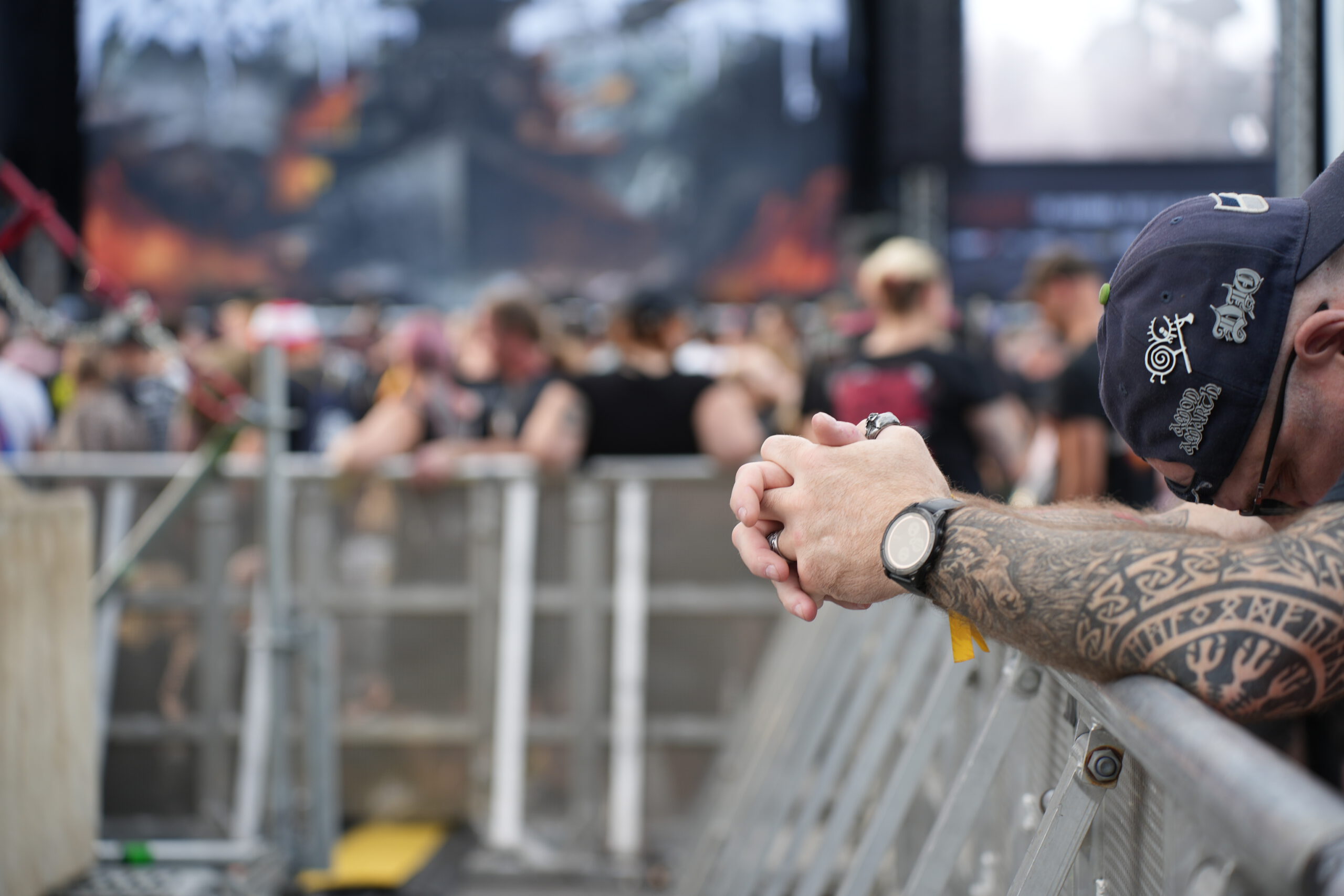 Man sieht die Arme eines erschöpften Fans an einer Absperrung lehnend. Im Hintergrund eine Festivalbühne.