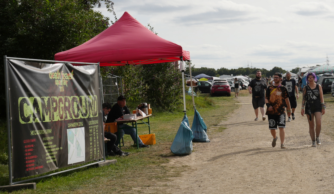 Blick auf den Eingangsbereich des Campgrounds des Death Feast Open Air in Andernach mit ein paar in Richtung Kamera laufenden Metalheads.